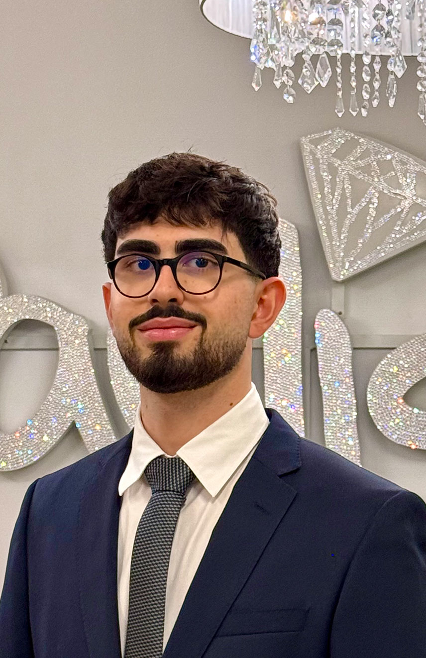 Ein Mann mit Brille lächelt sanft. Trägt Anzug und Krawatte, vor glitzerndem Hintergrund.
