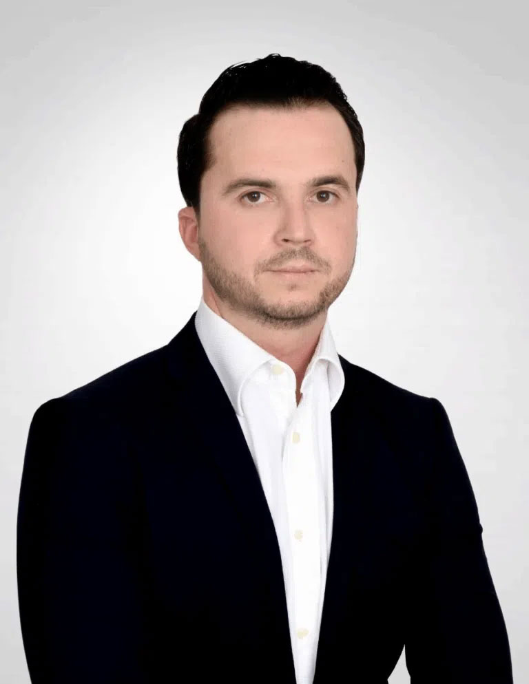 Eine ernst blickende Person im Anzug vor einem grauen Hintergrund.